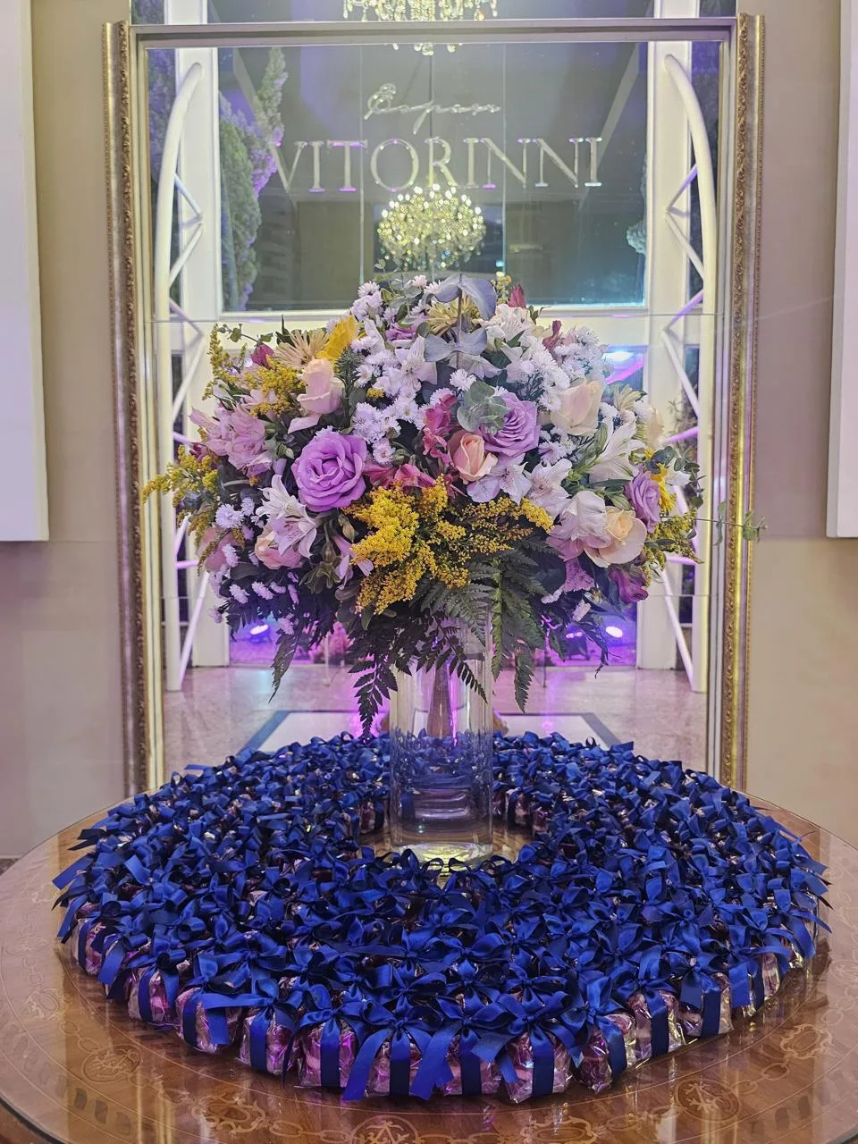 Centenas de bem casados embalados com fita e laço de cetim azul-marinho, em mesa com arranjo floral