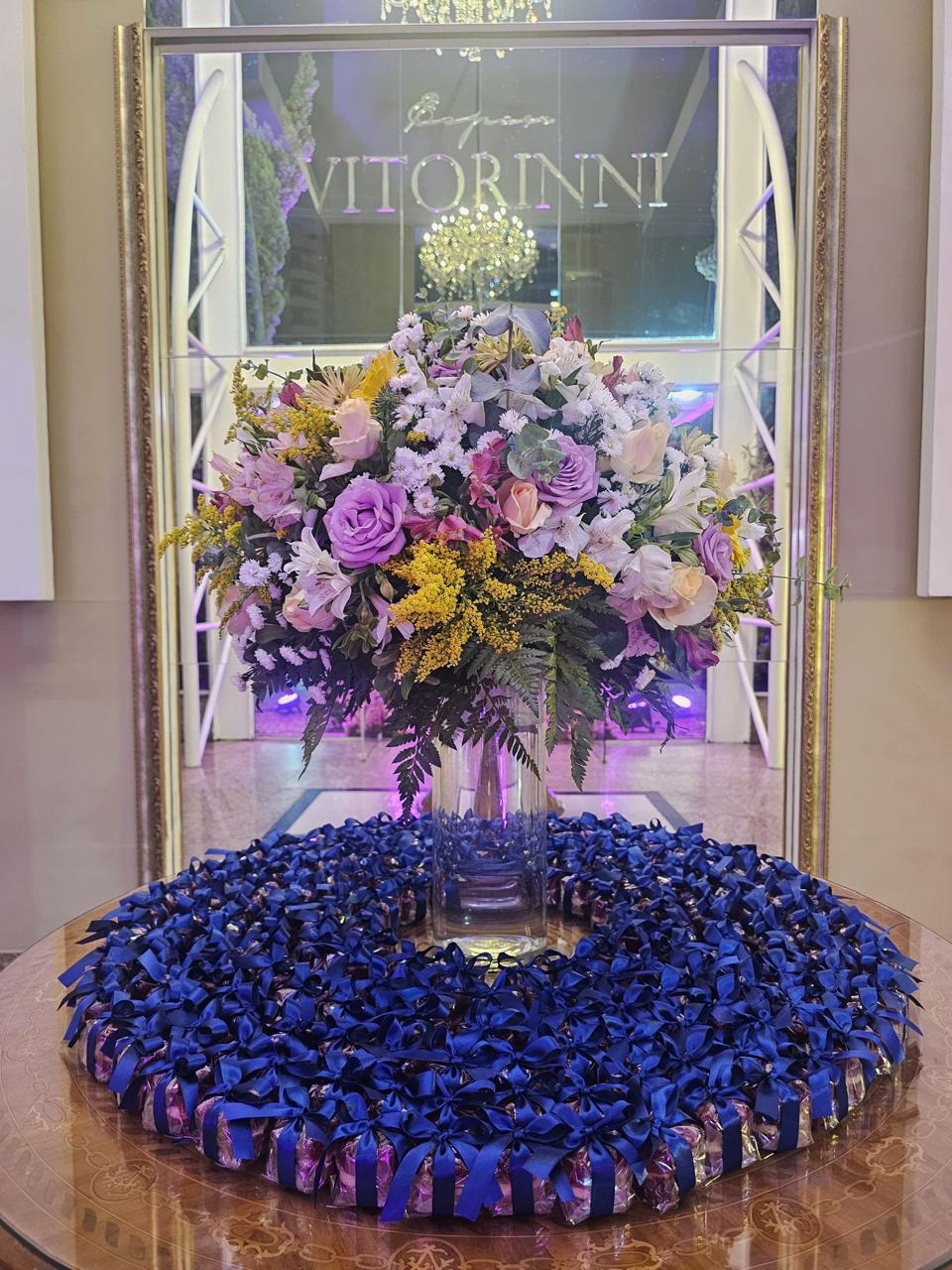 Centenas de bem casados embalados com fita e laço de cetim azul-marinho, em mesa com arranjo floral