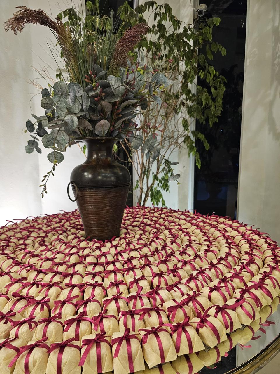 Bem casados em papel creme com fita e laço de cetim vinho, dispostos em espiral em mesa com folhagens