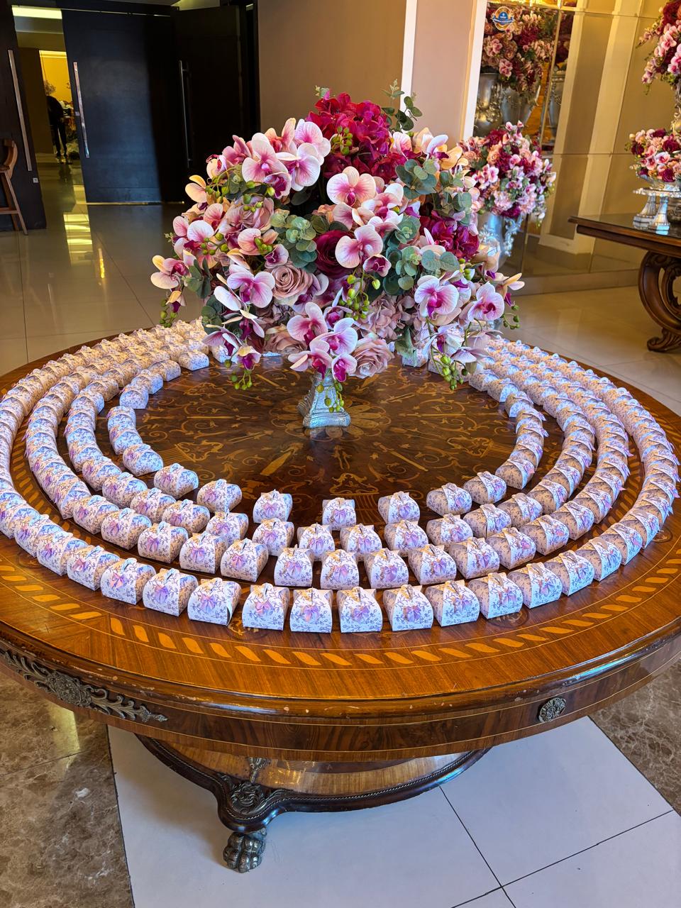 Caixinhas de lembrancinha com estampa floral rosa, dispostas em círculos sobre mesa com arranjo floral